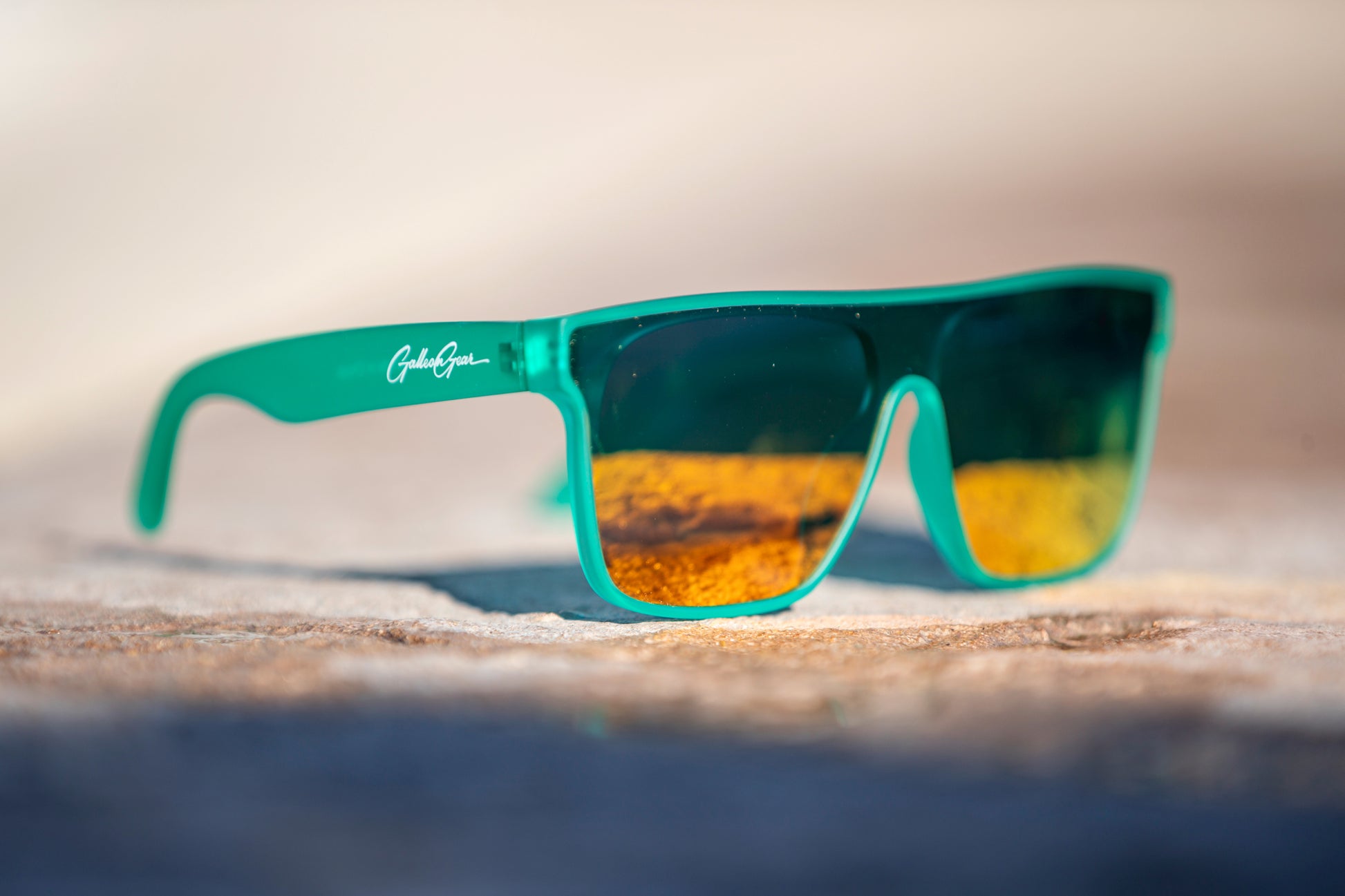 A bright colored sea foam green that is slightly transparent sunglasses with golden orange reflective lenses on a stone surface