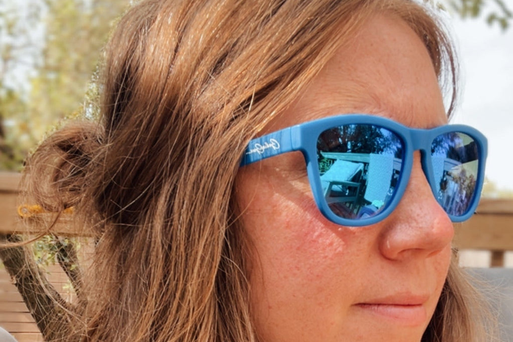A close-up shot of a woman with long light brown hair wearing blue framed sunglasses with blue mirror lenses, set against a blurred outdoor background of trees.