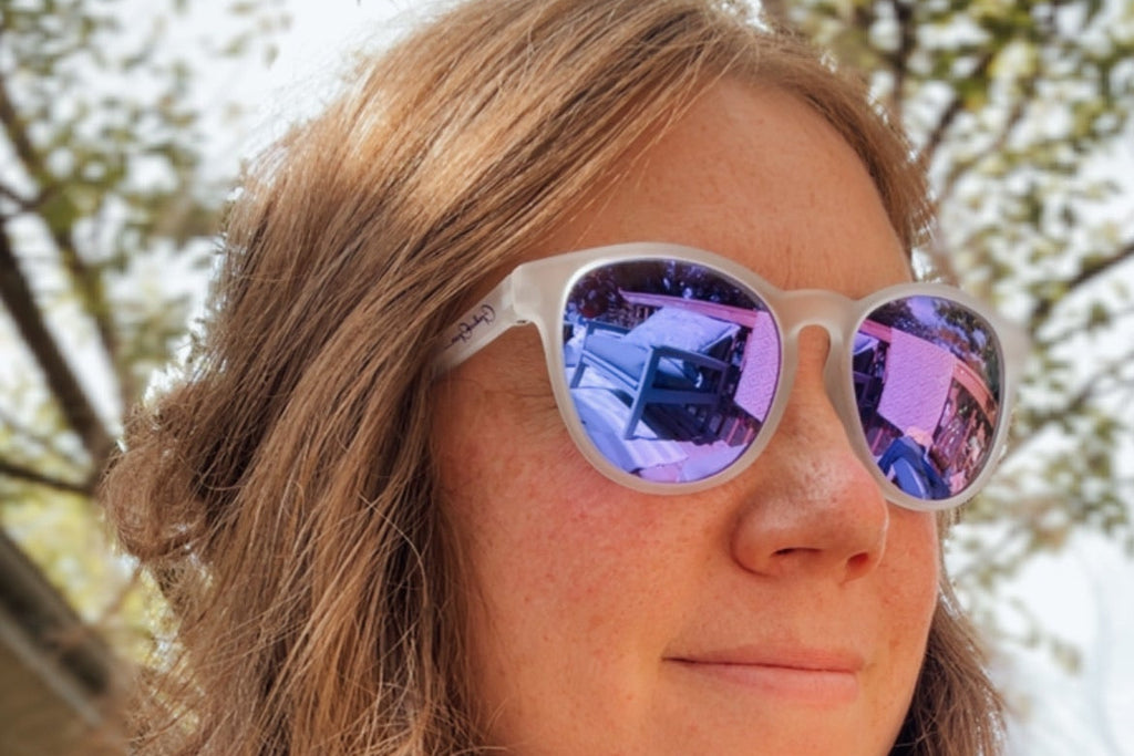 A close-up shot of a woman with long light brown hair wearing white-framed sunglasses with purple mirrored lenses, set against a blurred outdoor background of trees.