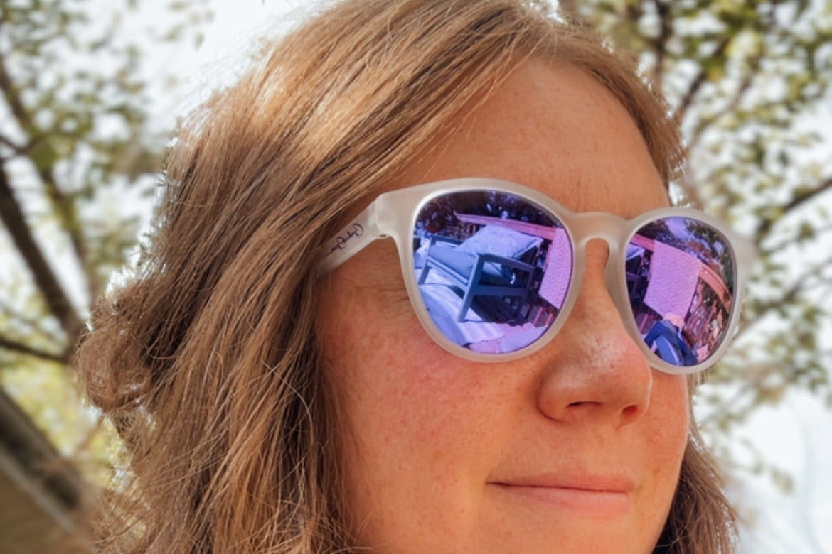 A close-up shot of a woman with long light brown hair wearing white-framed sunglasses with purple mirrored lenses, set against a blurred outdoor background of trees.