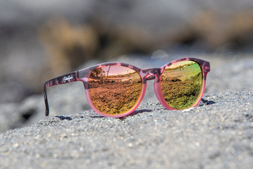 Blush pink with tortoiseshell frame sunglasses with rose gold/golden reflective lenses on a black sandy surface.