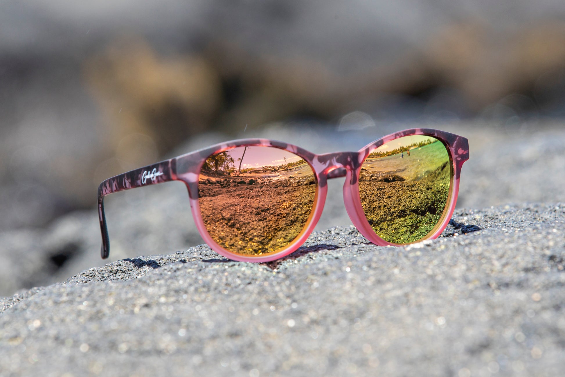 Blush pink with tortoiseshell frame sunglasses with rose gold/golden reflective lenses on a black sandy surface.