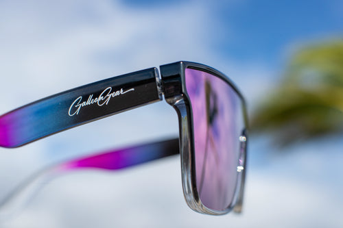 Sunglasses with a black frame with hints of slightly transparent pink and completely clear sections on the frame. The lenses reflect back a light purple.