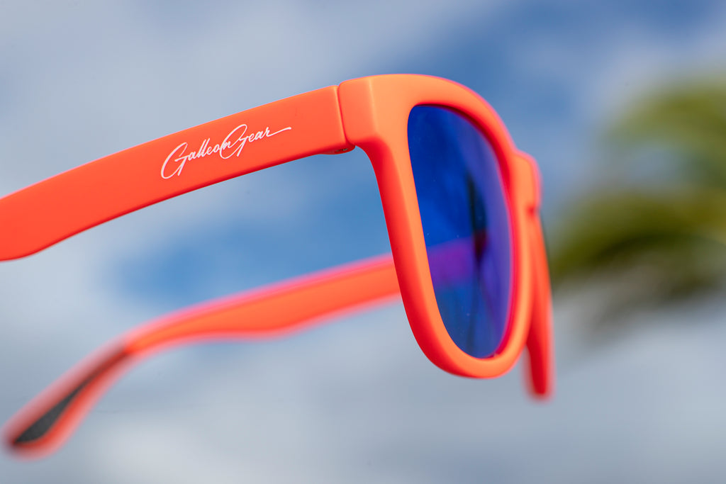 Close-up of brand logo on orange sunglasses with blue reflective lenses with a blurred sky and palm tree in the background.