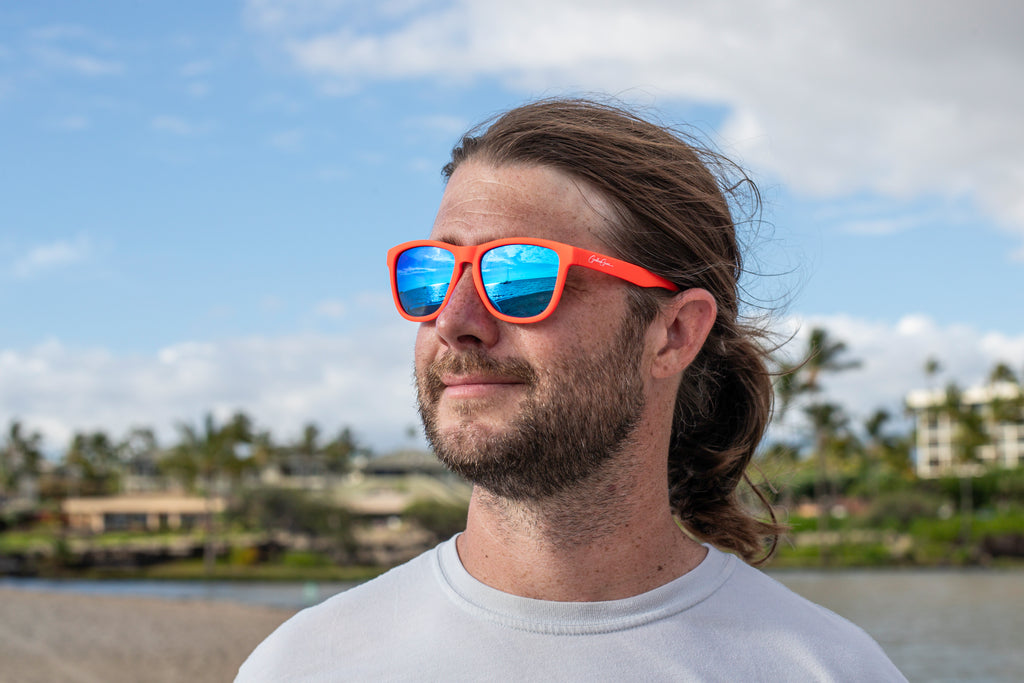 A man wearing orange sunglasses looking out at the ocean where a sailboat reflects on the blue sunglasses lenses.