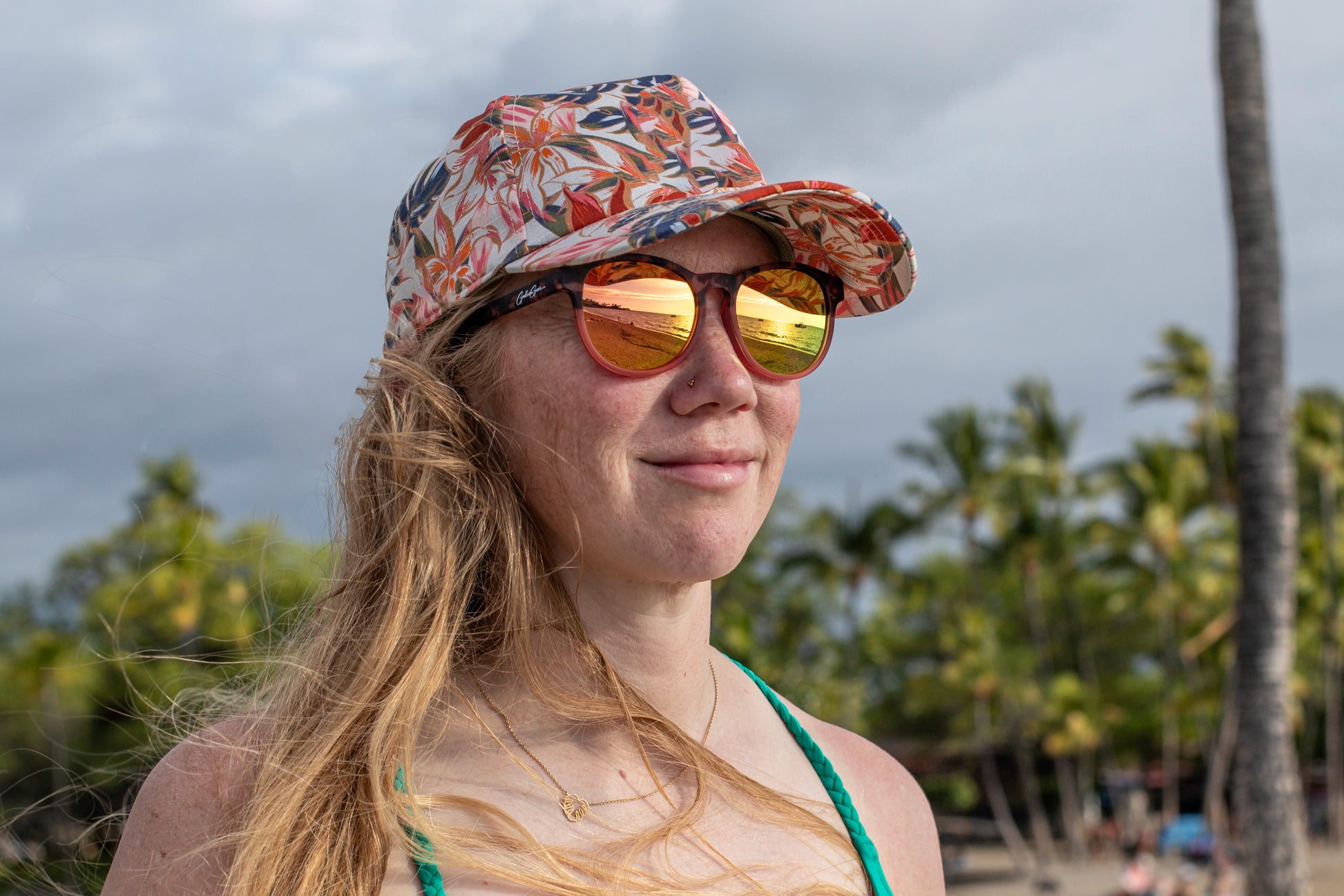 Woman wearing a floral cap and sunglasses reflecting the beach and ocean with palm trees blurred in the background