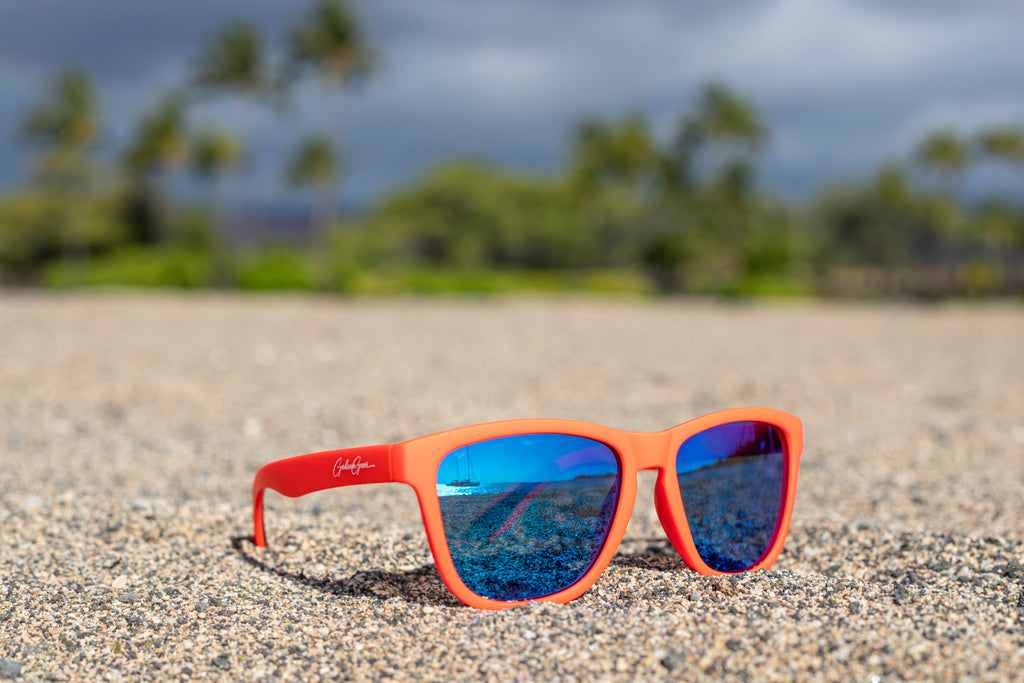 Orange sunglasses with blue reflective lenses on a sandy beach with blurred palm trees in the background. You can see a sailboat on the ocean in the reflection of the blue lenses.