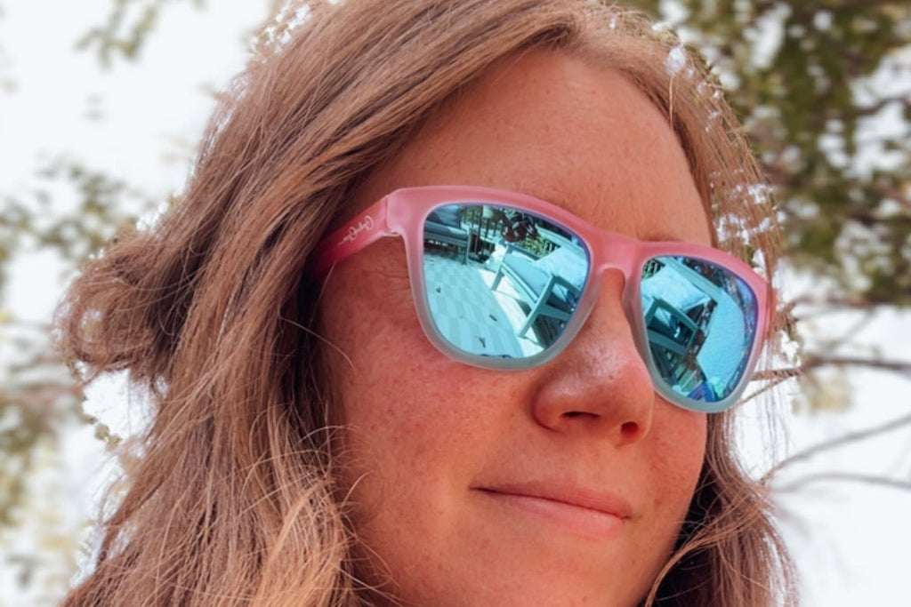 A close-up portrait of a woman with long light brown hair wearing pink-framed sunglasses with reflective blue lenses, photographed outdoors with blurred trees in the background.