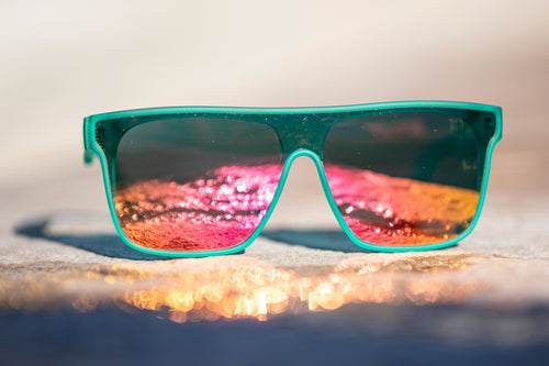 Sunglasses with a slightly transparent bright sea foam green frame and reflective pink and orange lenses on a blurred background