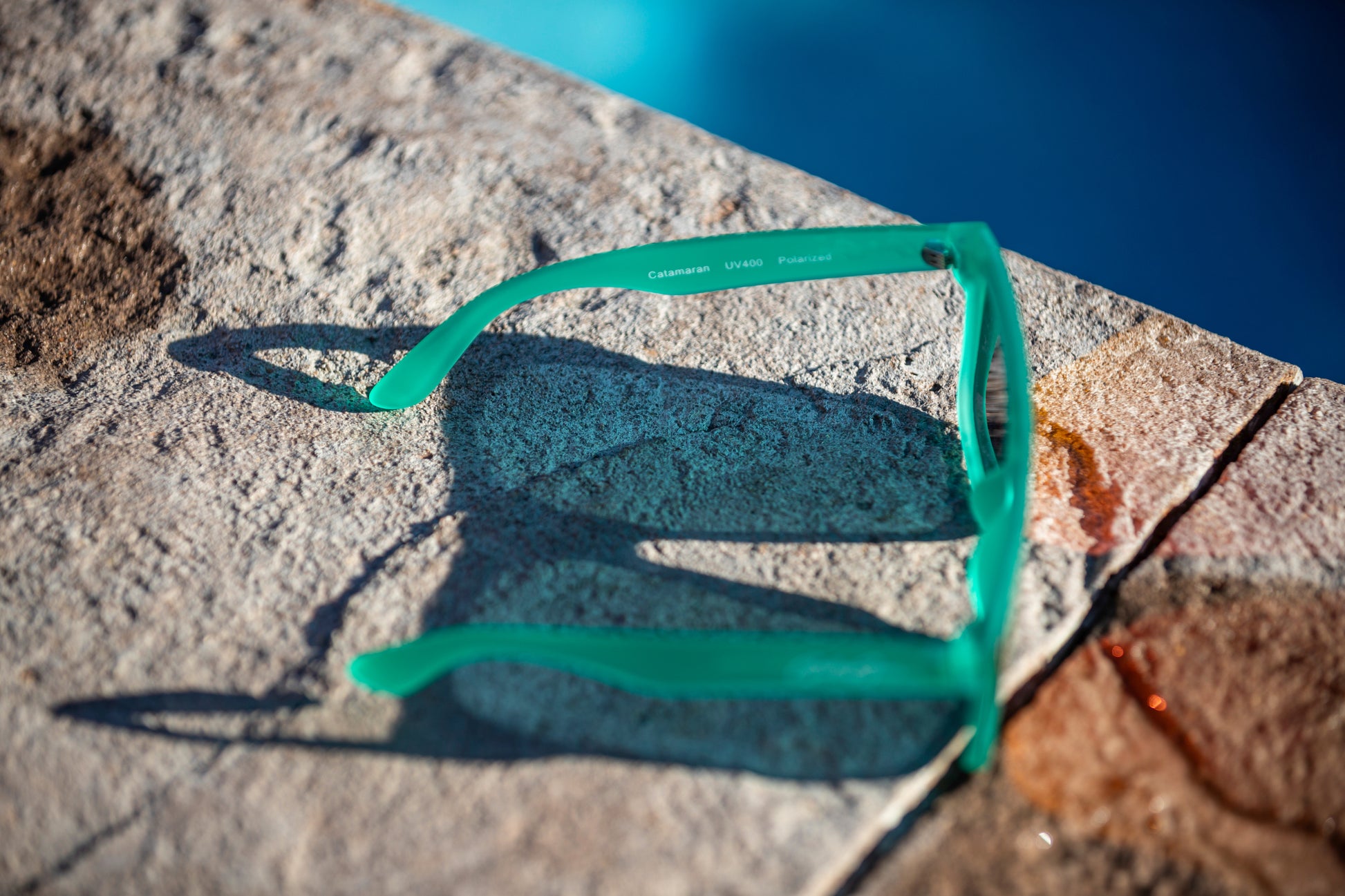 Slightly transparent bright sea foam green sunglasses on a stone ledge of a pool 