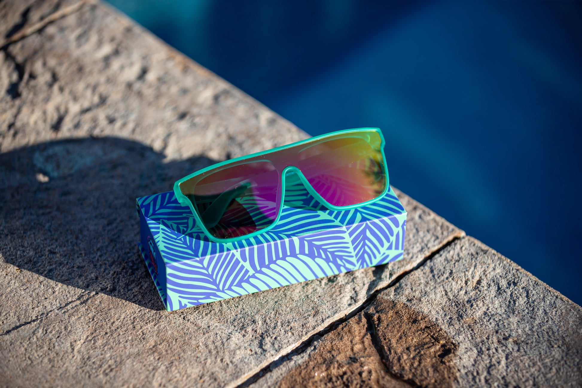 Sunglasses with colorful (pink/yellow/orange) reflective lenses on a patterned box by a poolside.