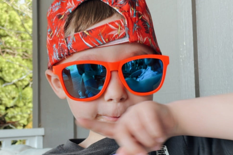 A young boy wearing bright orange-framed sunglasses with blue mirrored lenses and a red patterned cap, sitting on a porch. 