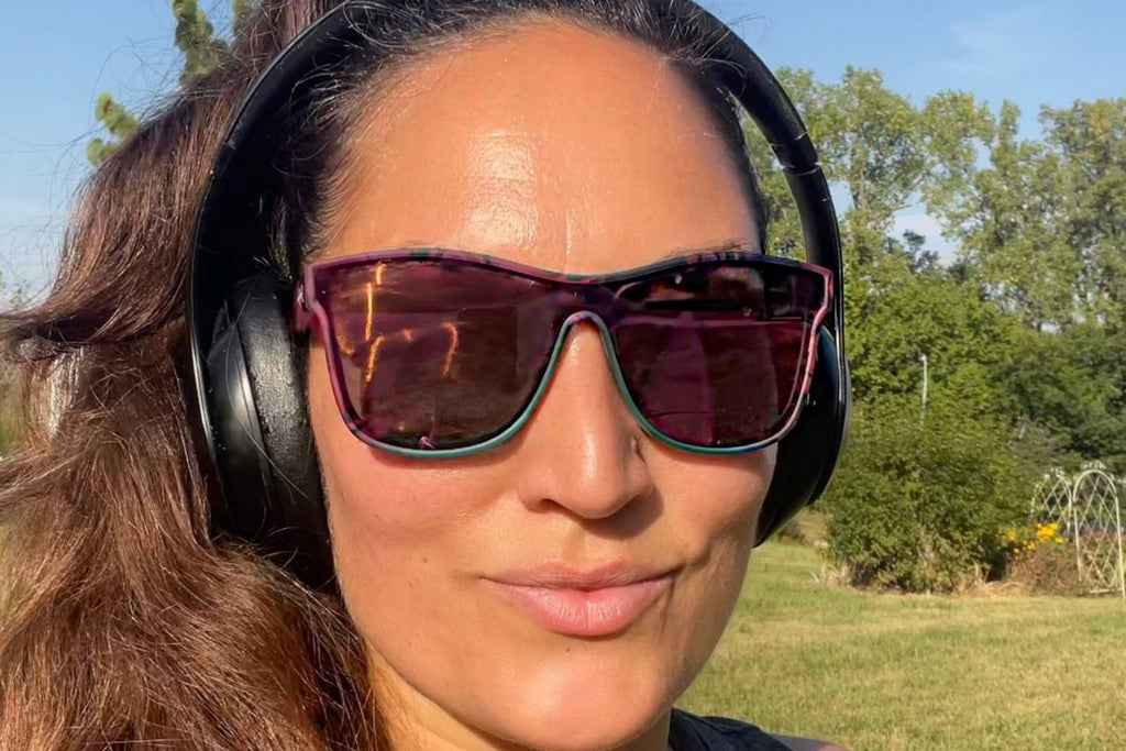 Woman wearing sunglasses and headphones outdoors with trees and sky in the background