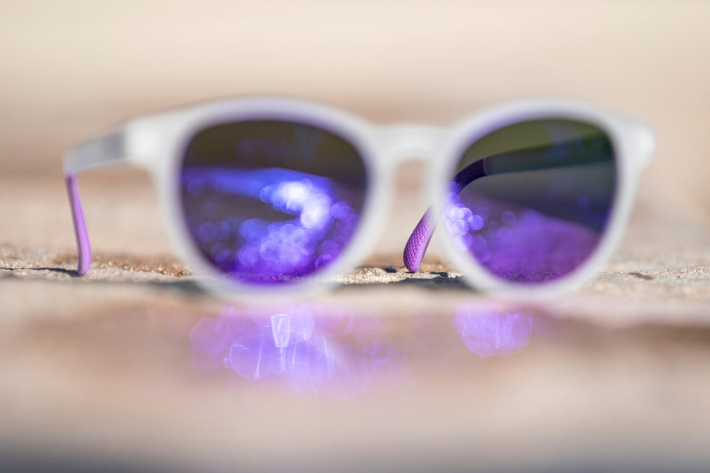 Blurred sunglasses with purple lenses with a focus on the purple temple grip on a blurred stone surface.