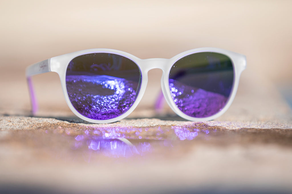 White semi-transparent sunglasses with purple-tinted lenses on reflecting sunlight off of a puddle.