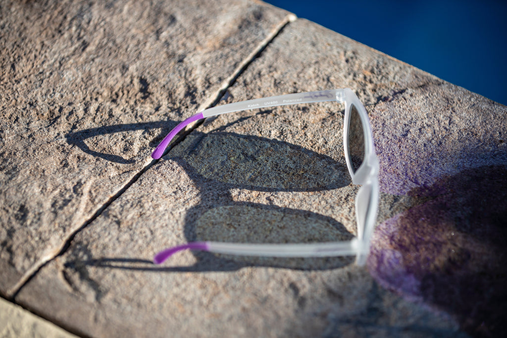 White semi-transparent sunglasses with a purple temple grips on a stone surface near water