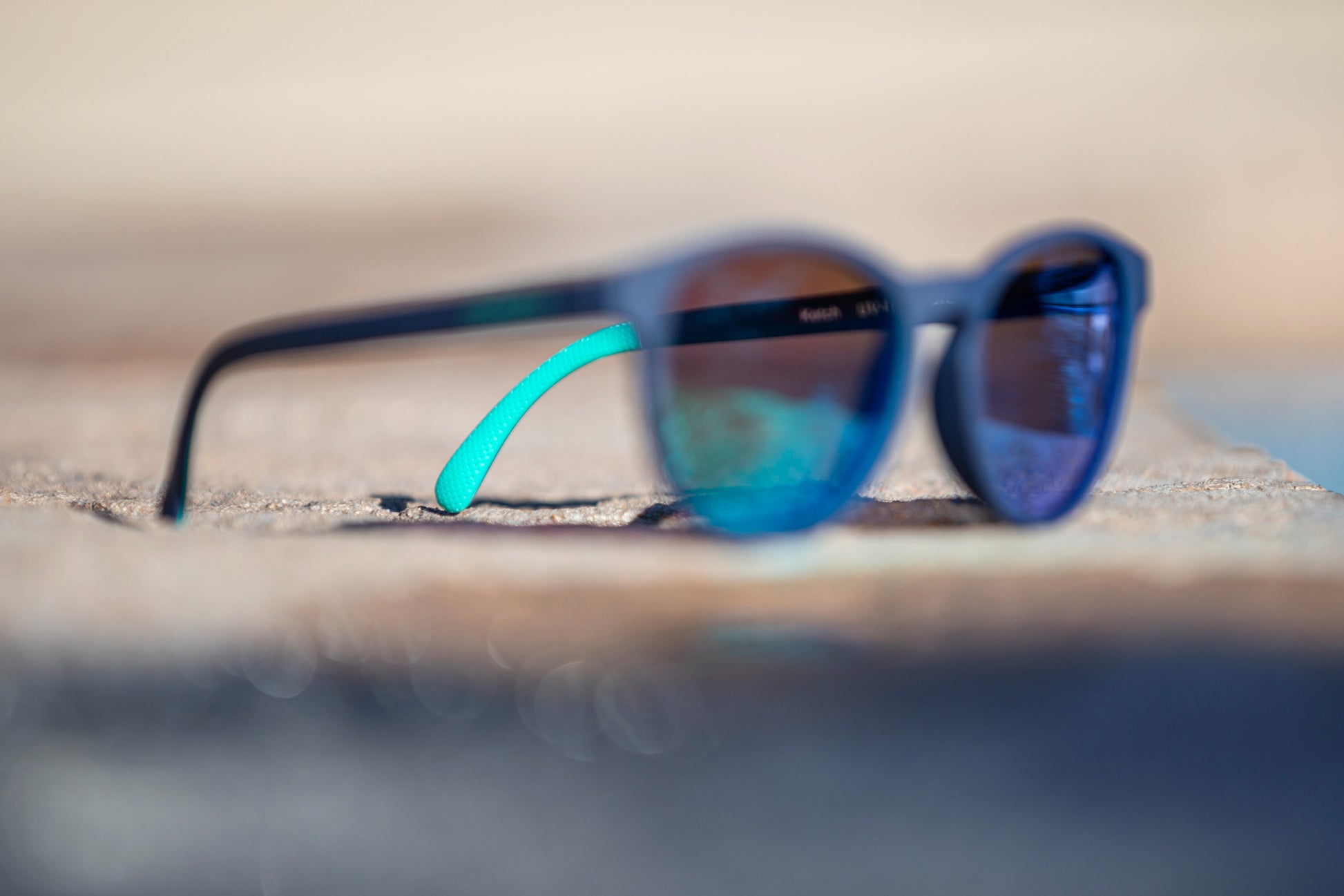 Blurred navy blue sunglasses on a textured surface with a blurred background with focus on the teal detail of the temple grip.