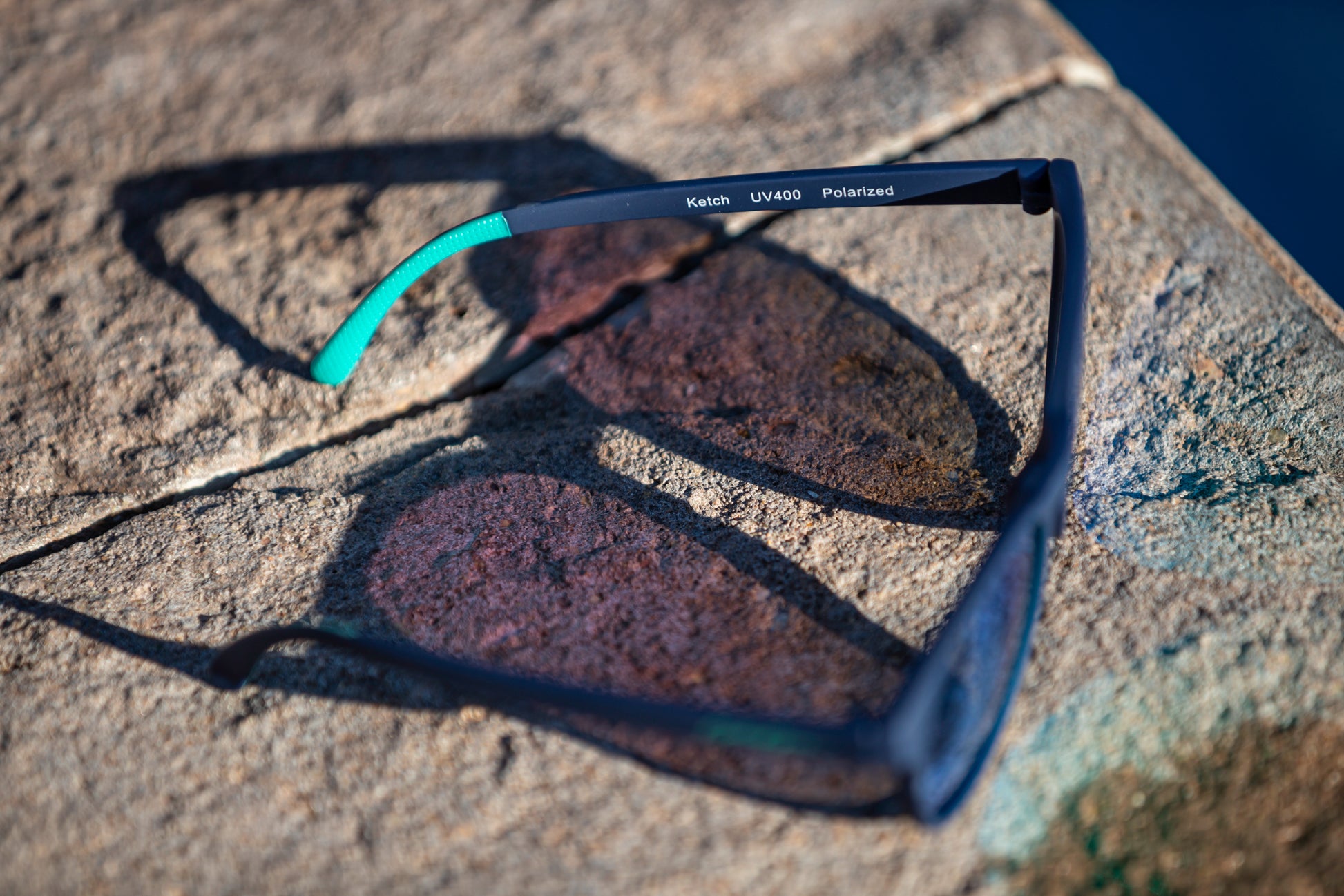 Sunglasses with a navy blue frame and teal temple grips on a textured stone surface next to a pool.