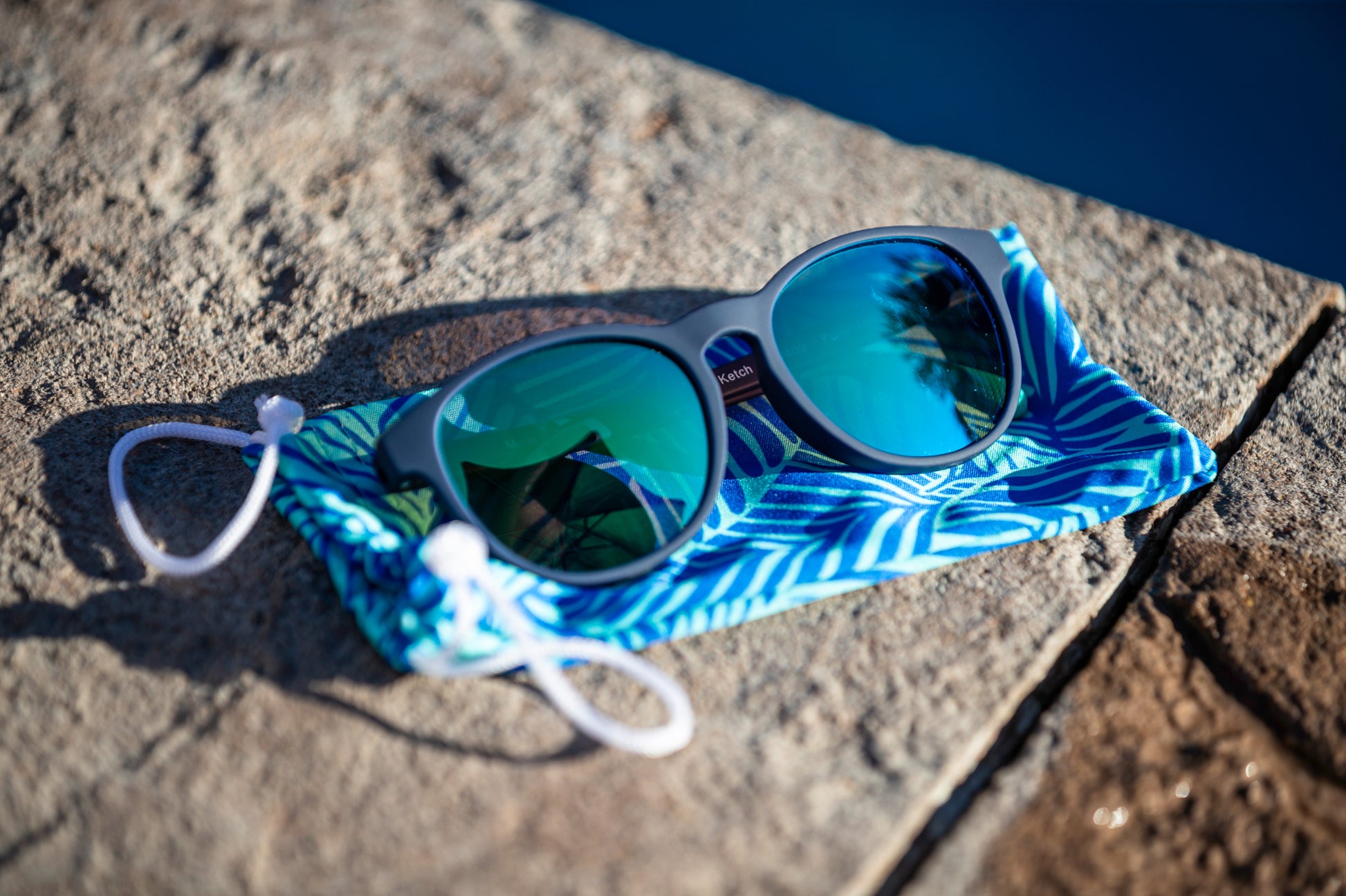 Navy blue sunglasses with a reflective surface on a blue patterned cloth bag, placed on a stone surface.