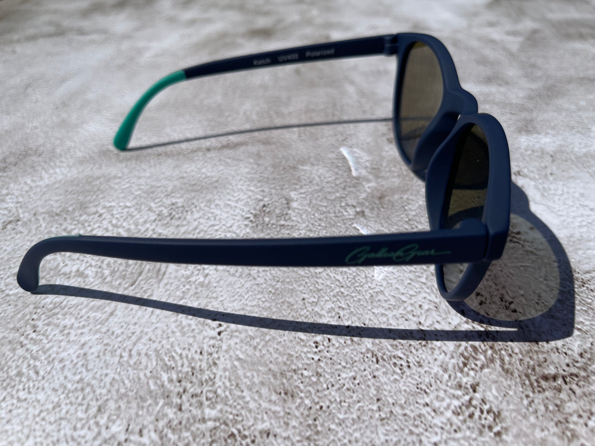 Navy blue sunglasses with teal temple grips on a concrete surface