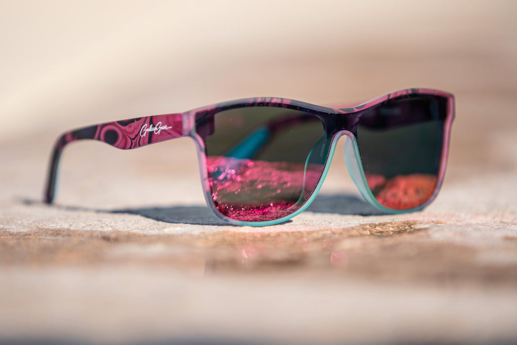 Sunglasses with pink and black frame with a hint of turquoise on the frame below the pink and orange reflective lenses on a blurred concrete background.