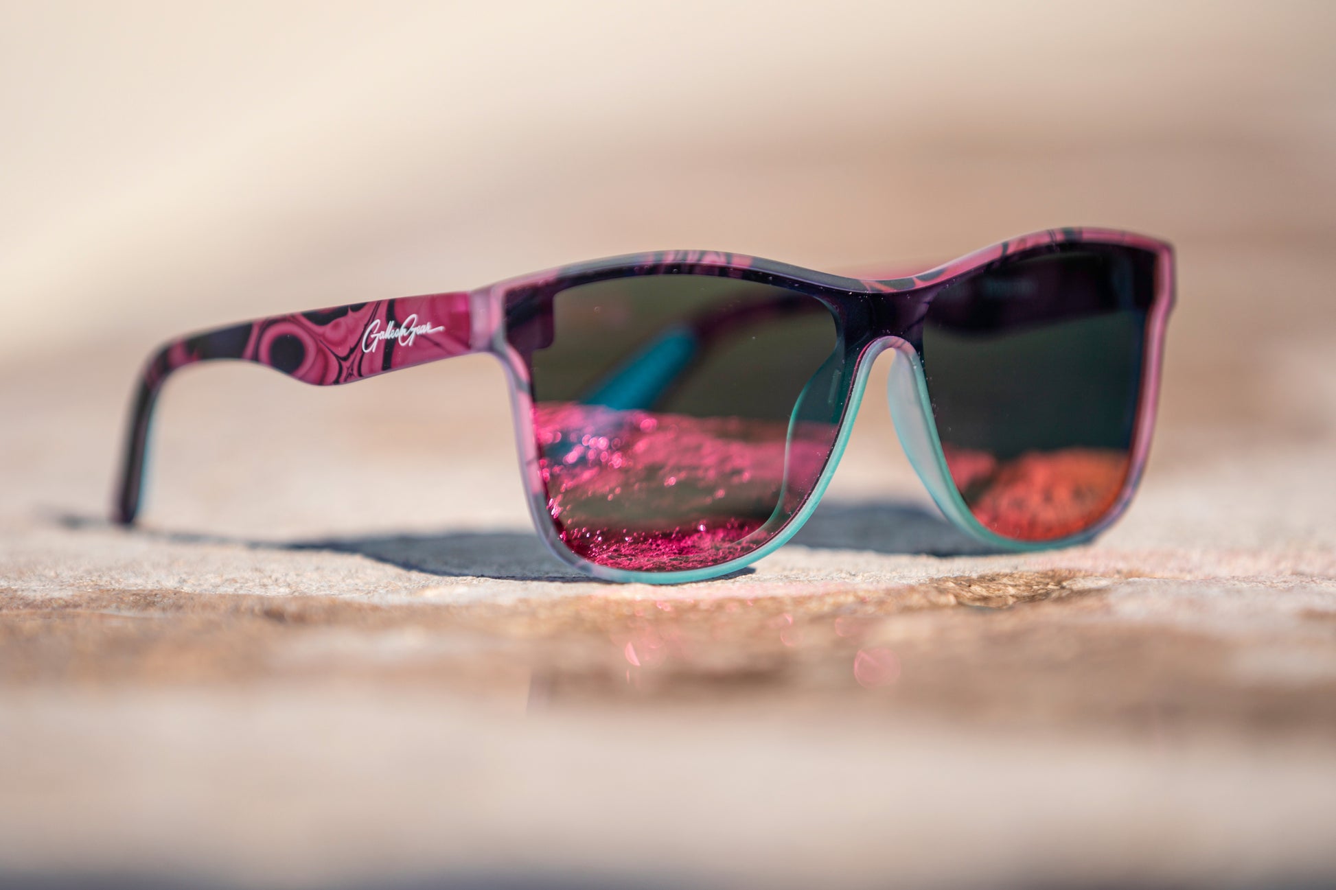 Sunglasses with pink and black frame with a hint of turquoise on the frame below the pink and orange reflective lenses on a blurred concrete background.