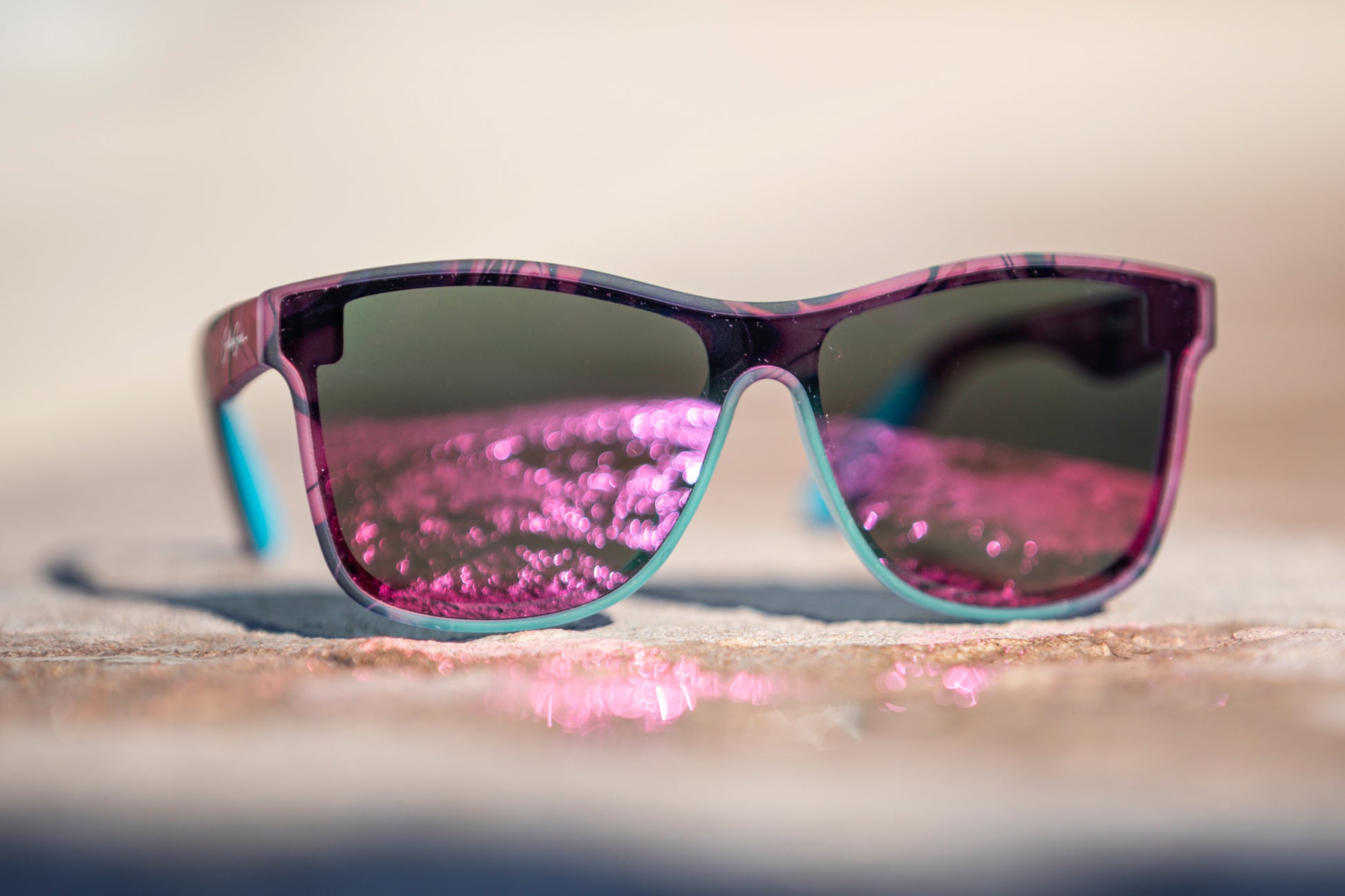 A pair of sunglasses with a pink, black, and turquoise frame with pink lenses reflecting the sunlight off of a puddle on concrete.