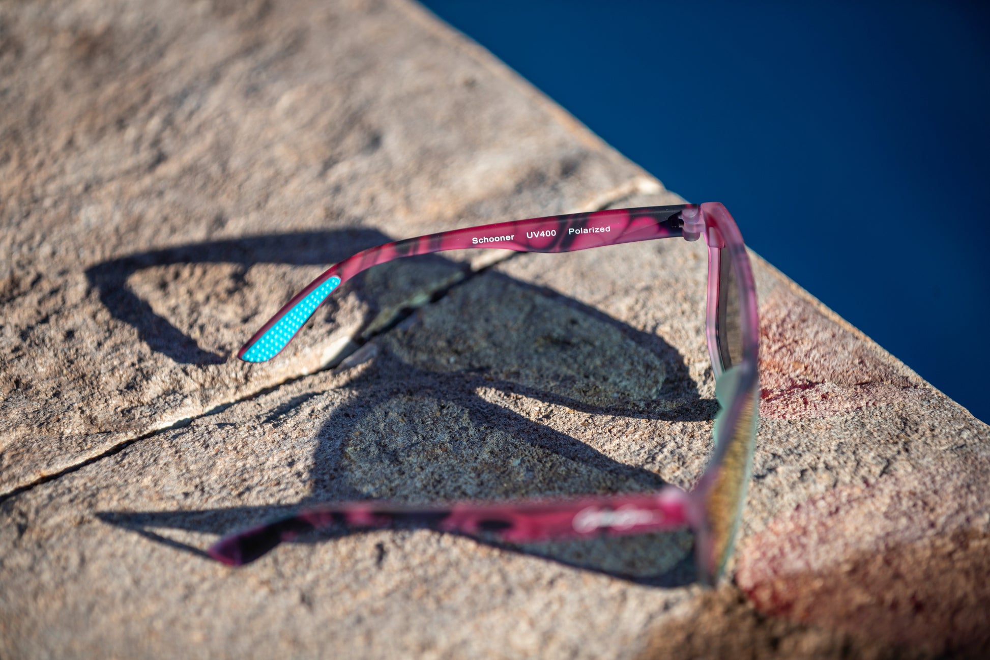 Sunglasses with a pink, black, and turquoise frame on a poolside stone surface with water in the background.