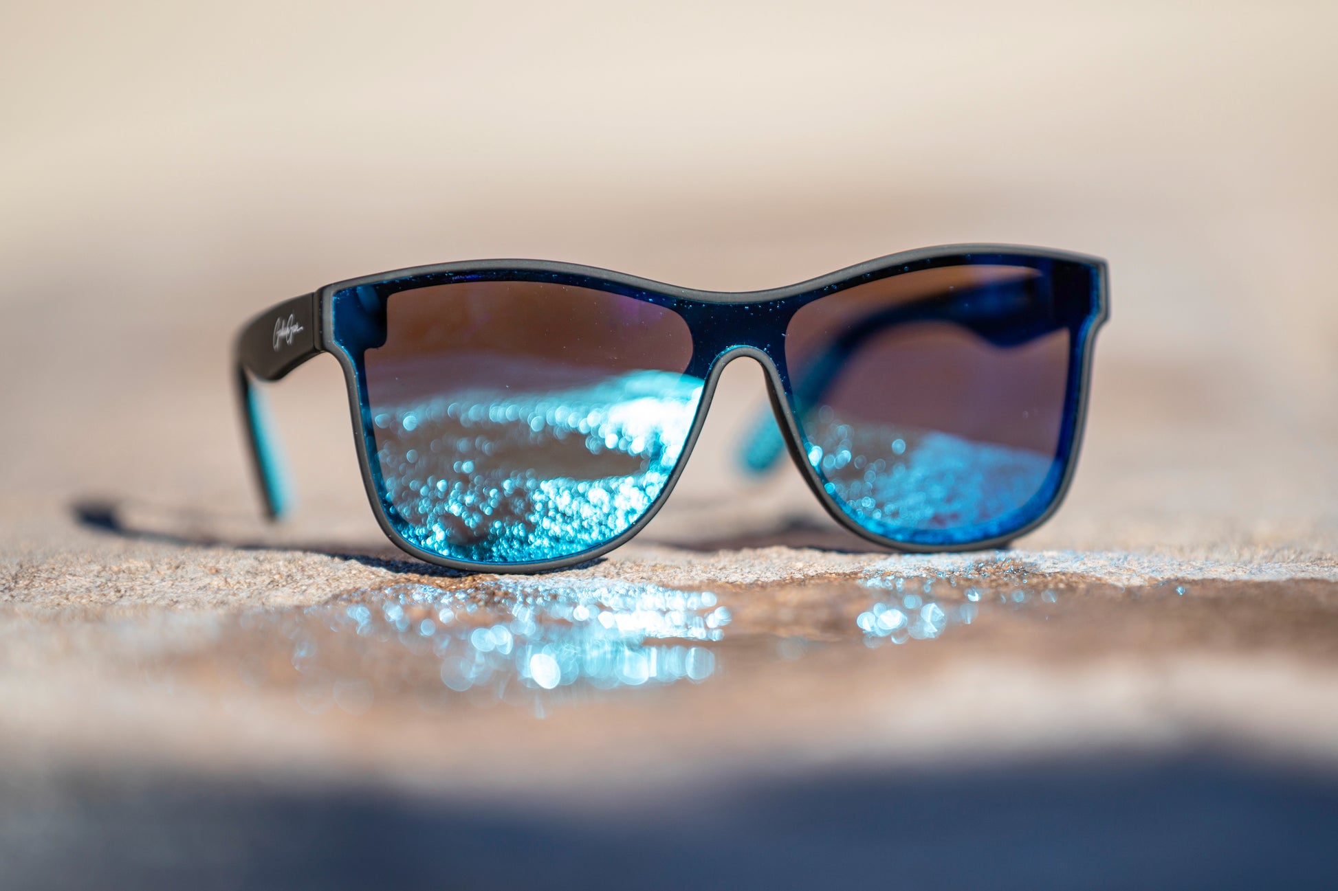 Black Sunglasses with a light blue reflection on the lenses of sunlight off of a puddle on a blurred stone surface.