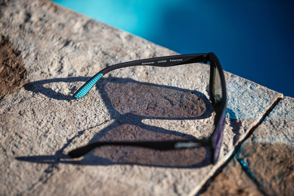 Black sunglasses with light blue temple grips on a on a stone surface near a pool.