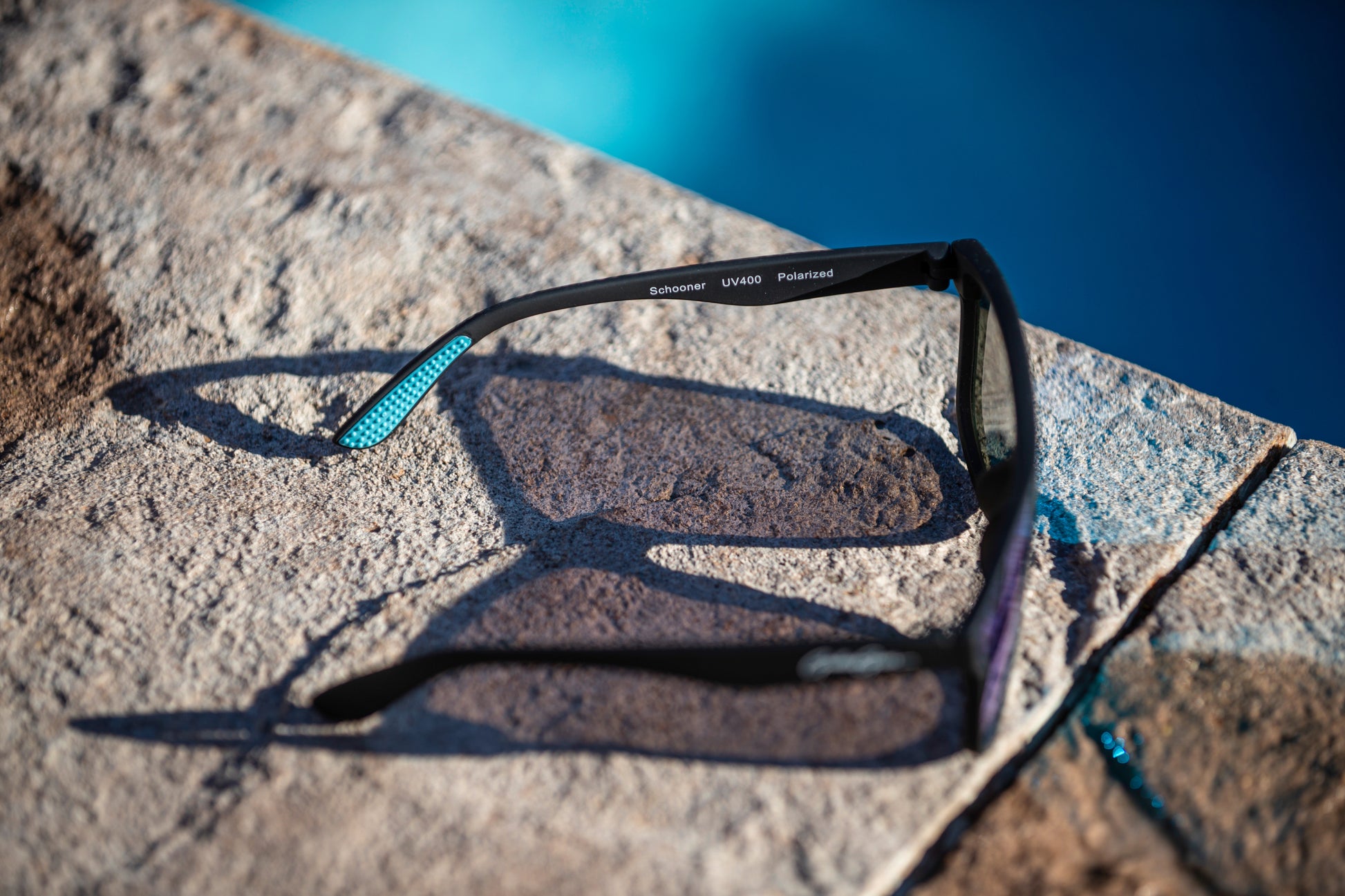 Black sunglasses with light blue temple grips on a on a stone surface near a pool.