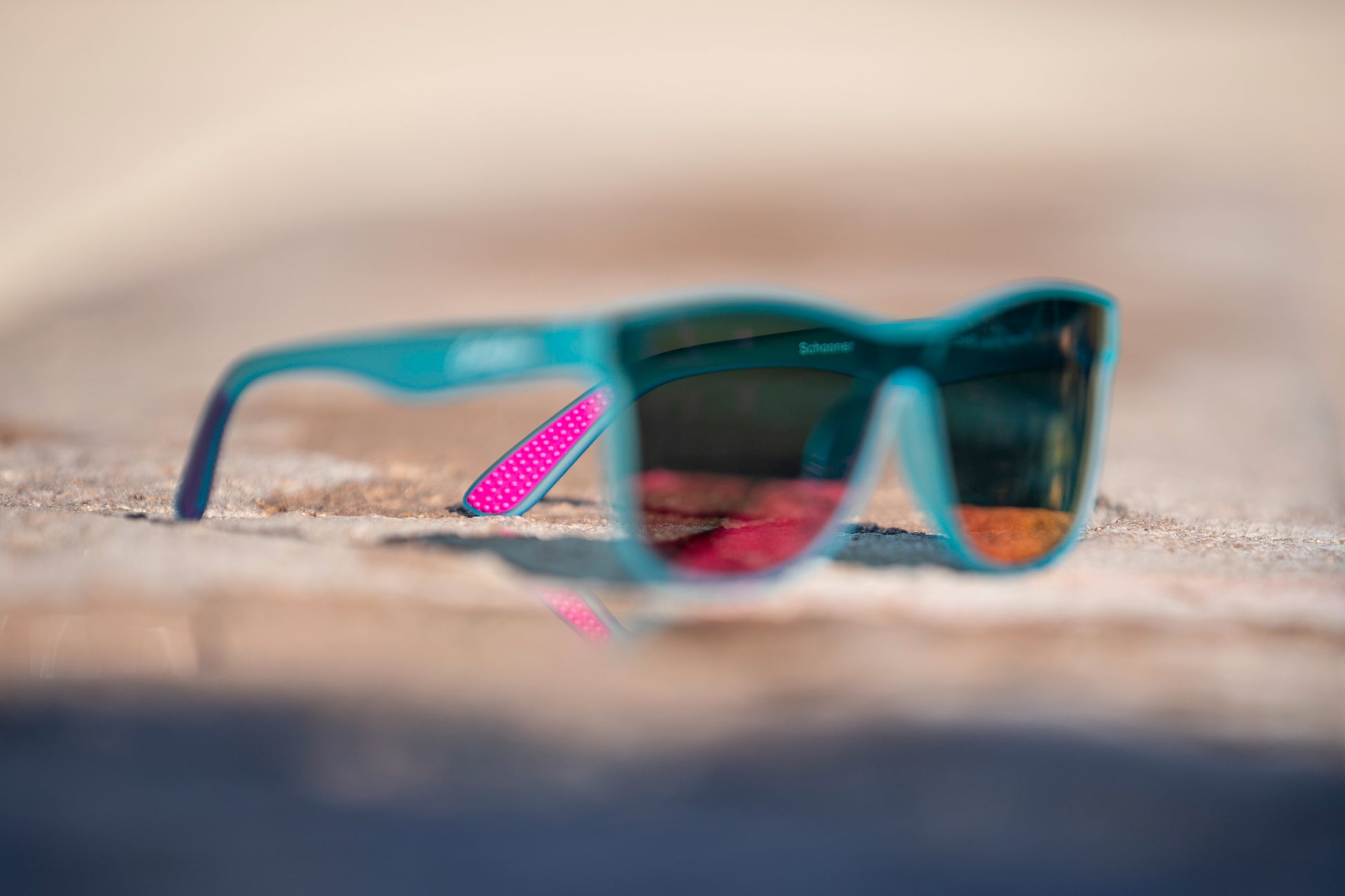 Blurred light aqua blue sunglasses with pink temple grip in focus on a blurred concrete surface.