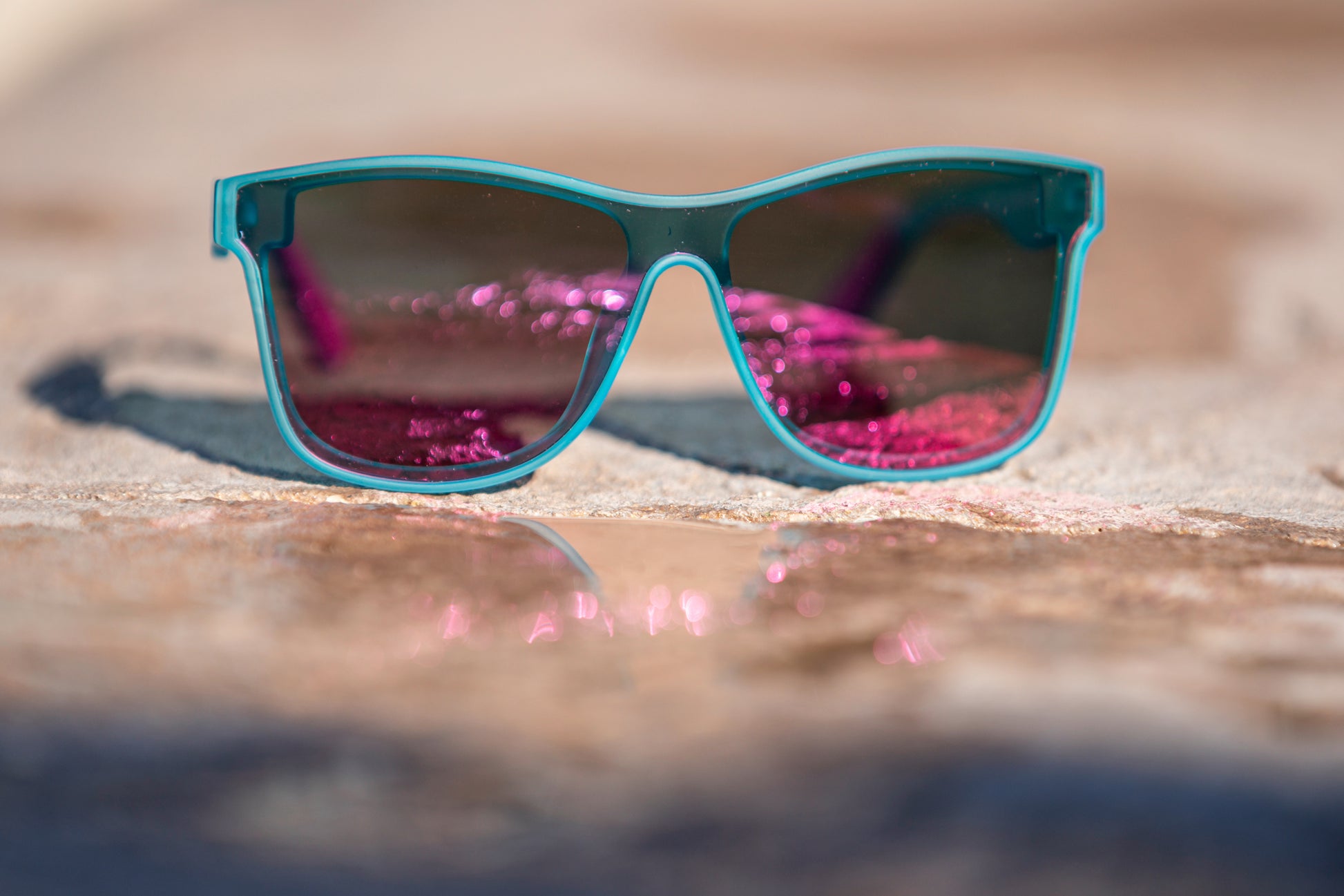 Sunglasses with a slightly transparent light aqua blue frame and dark pink reflective lenses on a concrete surface.