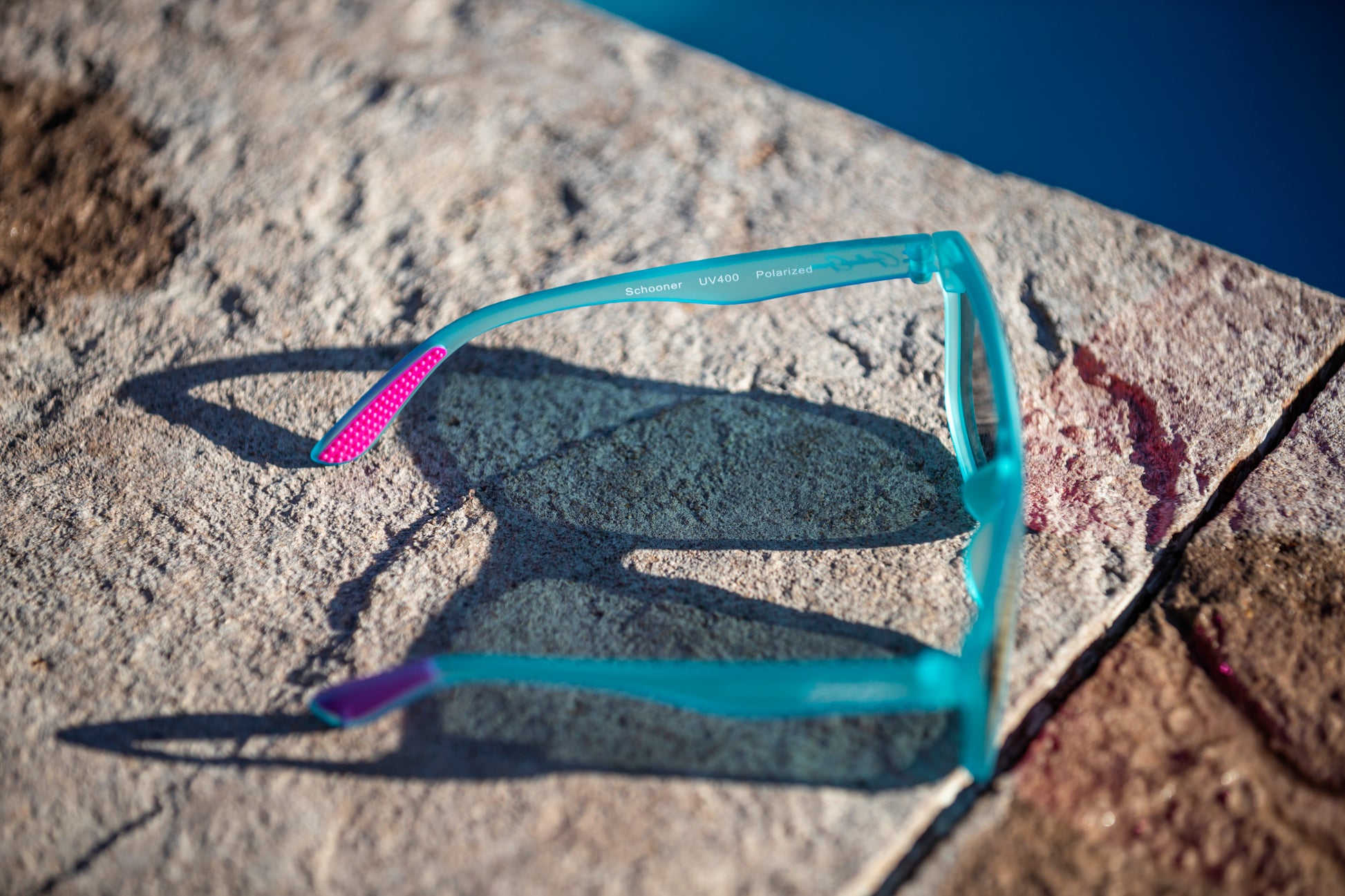 On overhead and side view of light aqua blue sunglasses with pink temple grips on a stone surface near water. The leg of the sunglasses says, "Schooner UV400 Polarized."