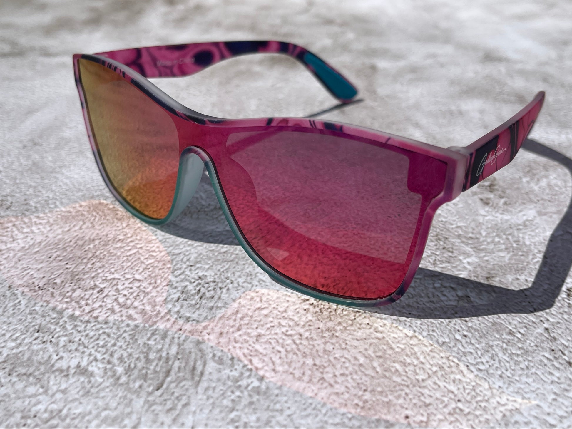 A pair of pink, black, and turquoise framed sunglasses with pink and orange reflective lenses on a textured surface,