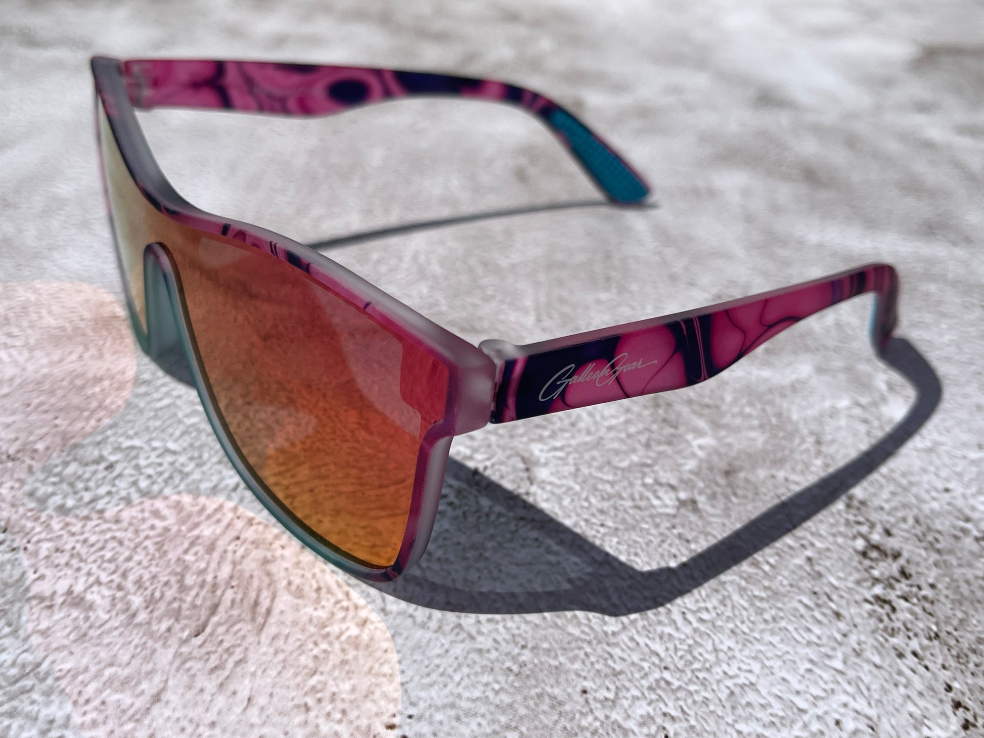 Pink sunglasses with a black marble design throughout the design and turquoise on the frame under the lenses that reflect back orange and pink on a textured surface.