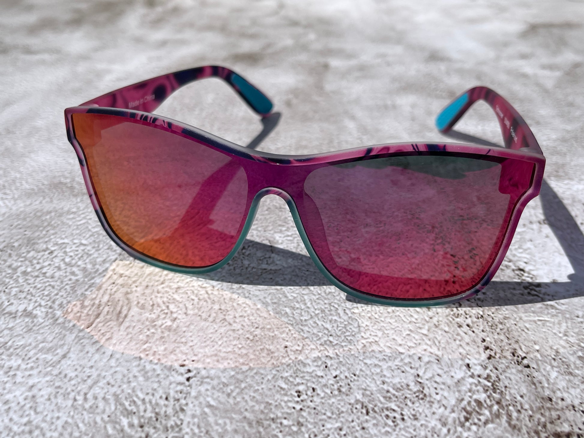 A pair of pink, black, and turquoise framed sunglasses with pink and orange reflective lenses on a textured surface.