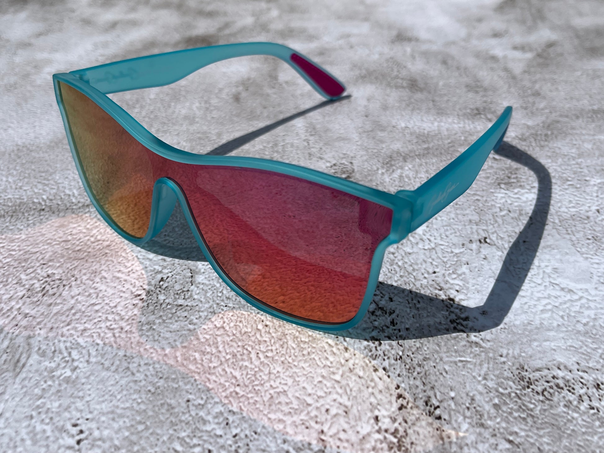 Sunglasses with light aqua blue almost teal frame and orange and red reflective lenses on a textured surface