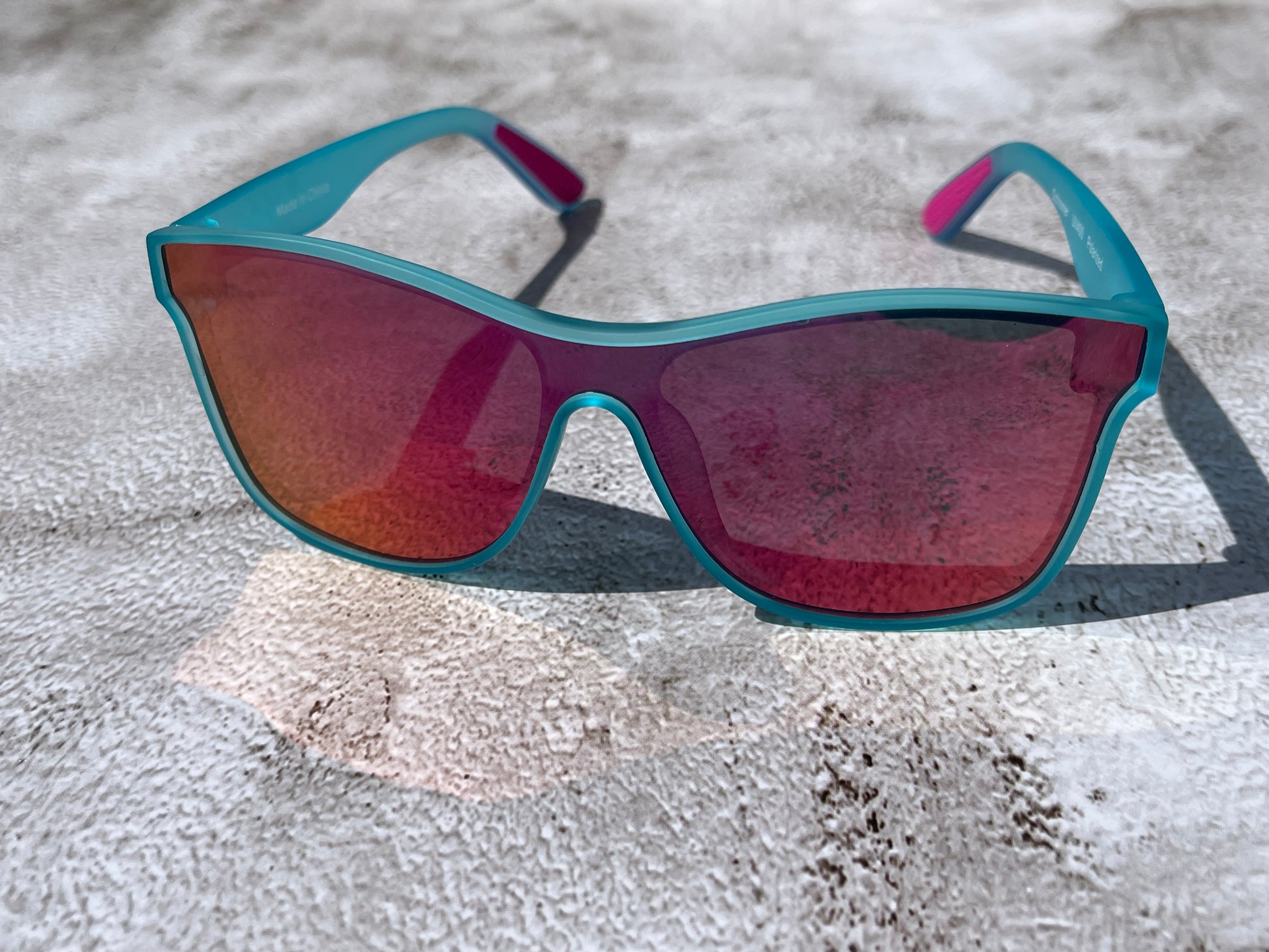 Sunglasses with slightly transparent light aqua blue almost teal frame and orange-pink reflective lenses on a textured surface