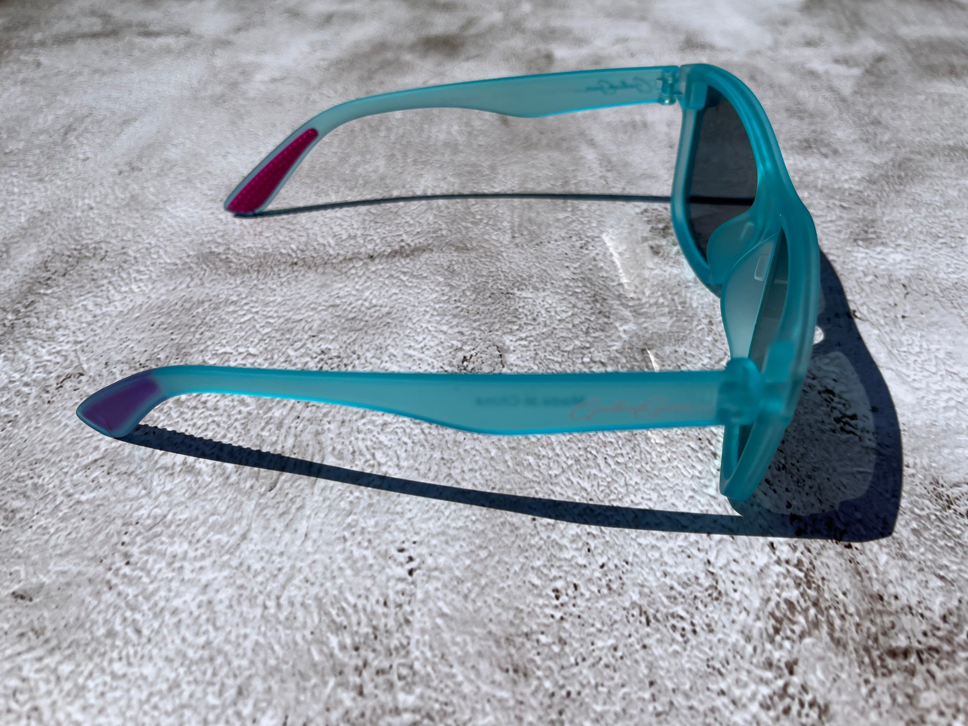 Slightly transparent aqua blue sunglasses  with pink temple grips on a textured surface.