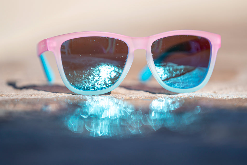 Pink and blue gradient sunglasses with blue lenses reflecting the sunlight through a puddle on concrete.
