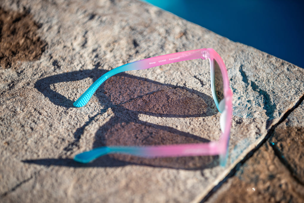 An overhead and side view of pink and blue gradient sunglasses on a pool's edge, and the sunglasses say Sloop UV400 Polarized on the inner left leg.