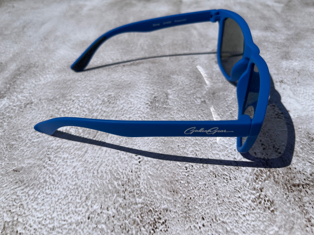 A side view of blue sunglasses with a brand logo on a concrete surface.