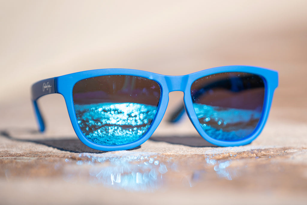 Blue sunglasses with blue lenses reflecting the sunlight off of a puddle on the concrete.