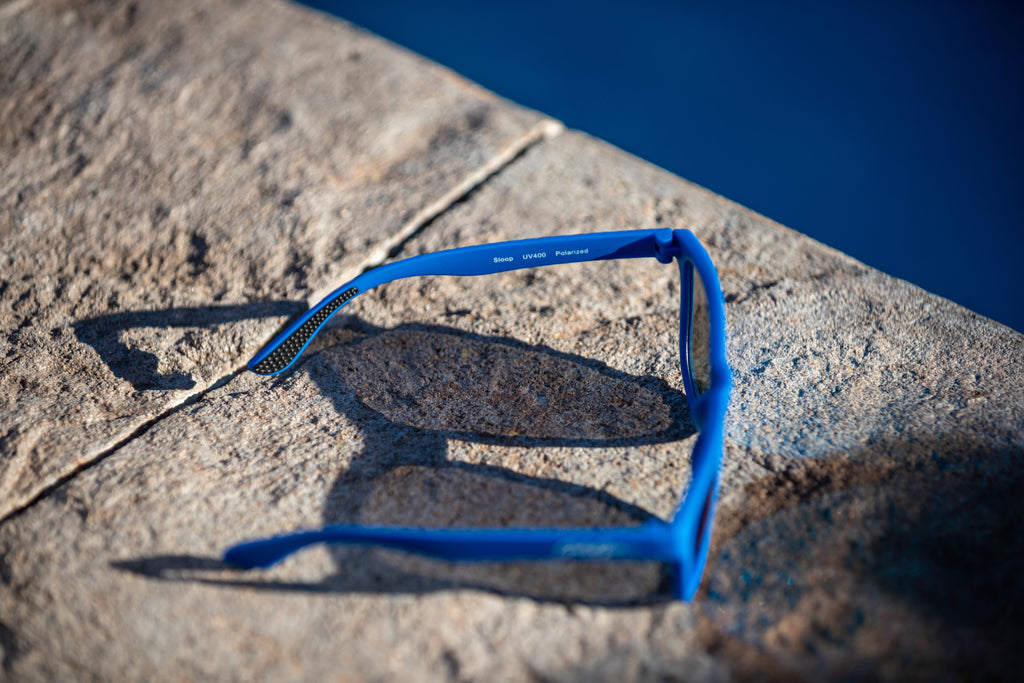 An overhead and side view of a pair of blue sunglasses on a pool's edge. It says Sloop UV400 Polarized on the inner left leg.